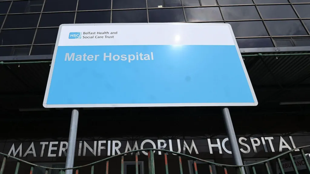 A blue hospital sign at the front of the Mater Hospital in Belfast. The tall building has a dark glass facade. The entrance also has a sign that says: "Mater Infirmorum Hospital" in large silver lettering. The blue sign, erected by the Belfast Health and Social Care Trust, is behind a railing with chipped green paint.