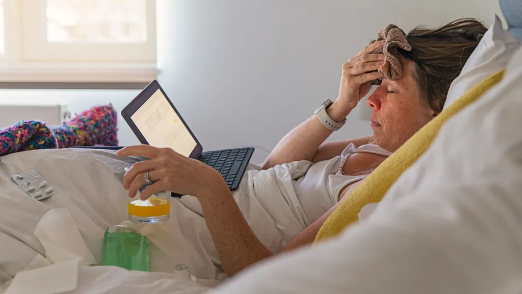 An ill woman lies in bed with her laptop. She has a flannel on her head and is drinking a glass of orange juice. 