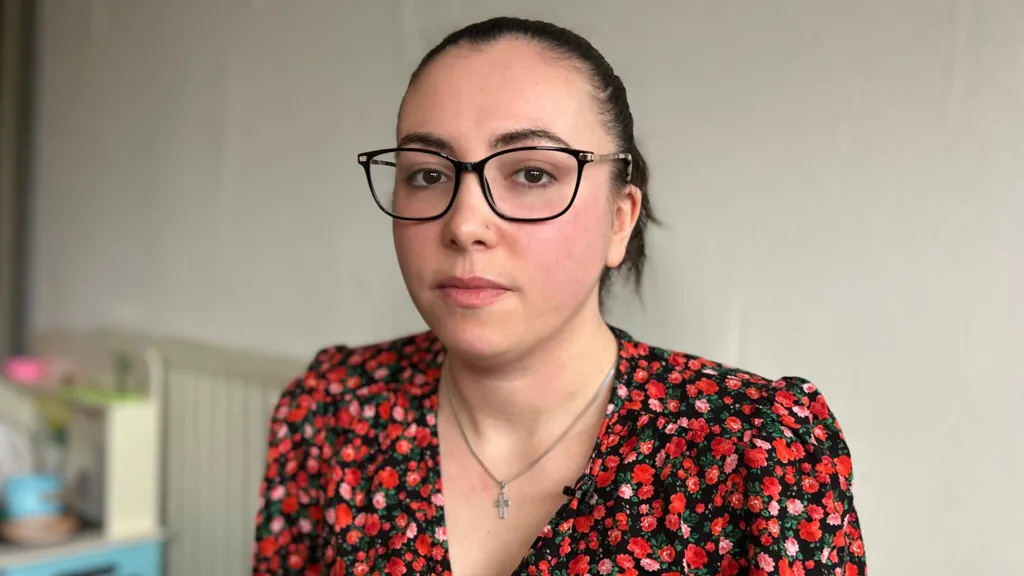 A woman sitting in her living room stares into the camera lens. She has brown hair scraped back in a ponytail, and is wearing a floral red top, a cross necklace, and black-rimmed glasses. 