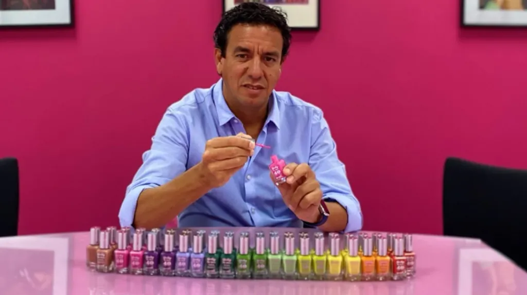 Man with dark hair and brown eyes, wearing a mid-blue button-up shirt, sits at a table on which there is an array of nail vanishes in different colours including pink, purple, lilac, green, yellow and orange polishes. 