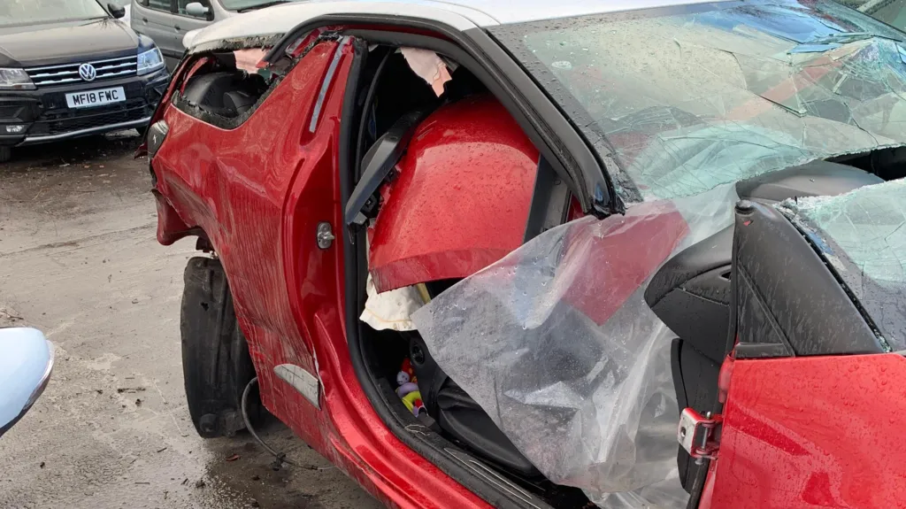 Image shows a red Citroën sat amongst other cars. It shows the damaged after the accident Rebecca was involved in. There is no drivers side door left on the car. The windscreen is smashed and has been cut in two. There are a number of car parts which have been piled on the drivers side seat. The back wheel is at an angle and the tyre is missing. 