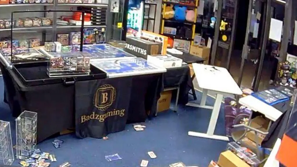 A CCTV image showing Badzgaming, a gaming shop after it had been broken into. There is stock, mainly cards, scattered over the blue floor and items are damaged. The glass door is open, with a white tale askew by the entrance. 