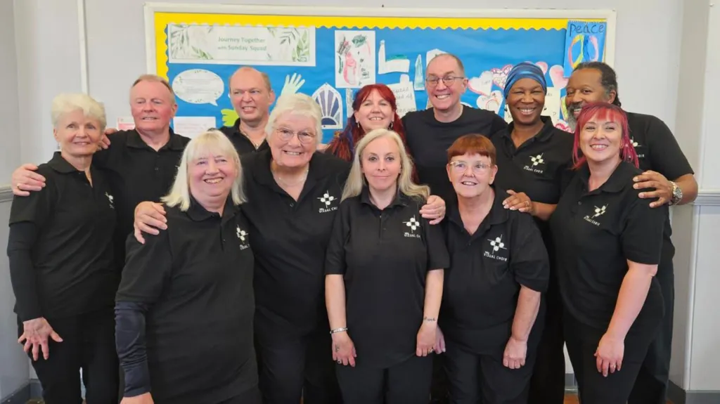 A group of 12 people stand shoulder to shoulder in two rows in a white-walled room and smile at the camera. They are made up of seven women and four men, all wearing black polo shirts with white logos. Behind the group is a blue and yellow picture board with hand-drawn pictures and artwork.