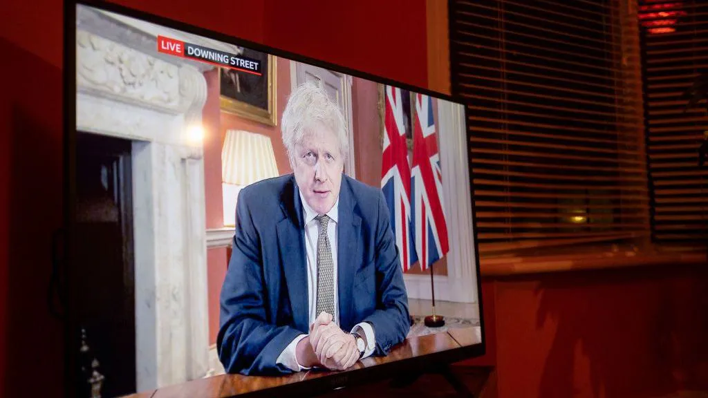 A photo of a TV screen in a living room shows Boris Johnson giving a briefing live from Downing Street