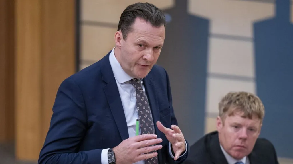 Russell Findlay, who has  dark hair slicked back, speaks with his hands held out in front of him. He is wearing a blue suit and shirt with a patterned tie. 