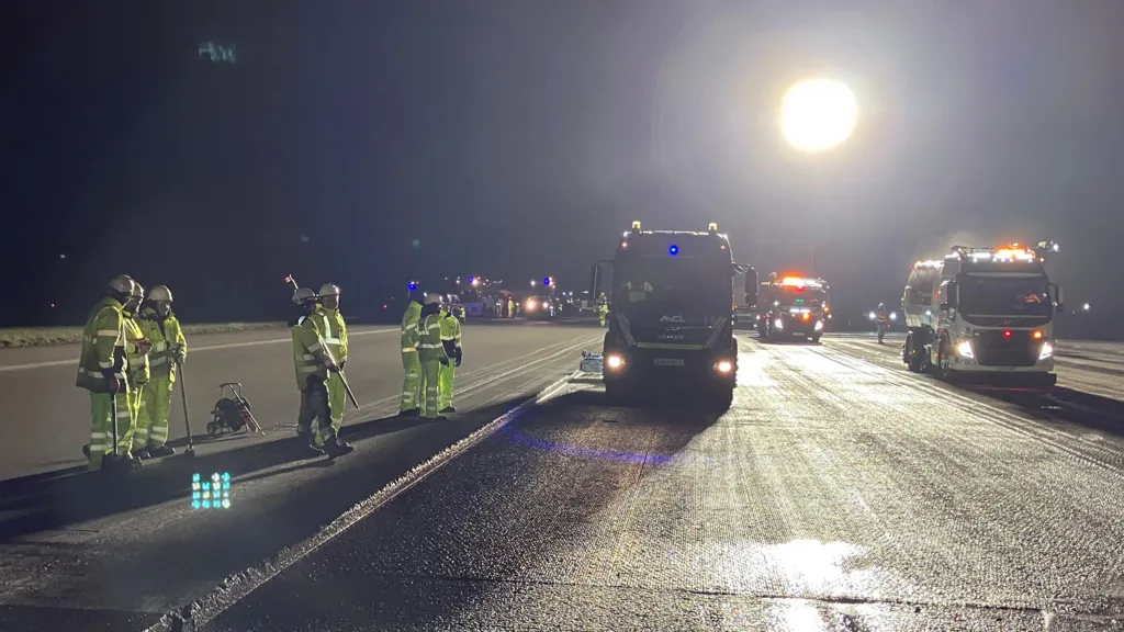 The image shows a well-lit airport runway at night with a large floodlight at the back of the picture. There are three large construction vehicles in the foreground with a group of workers in high-visibility clothing standing to the left side of the picture. There are a series of small lights in the distance. 