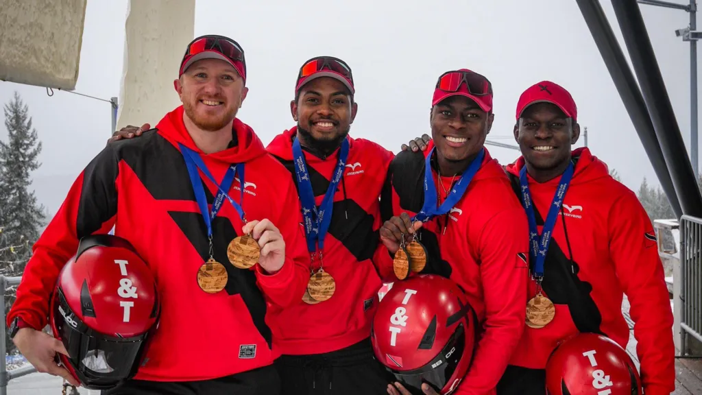 Four Trinidad and Tobago athletes in red helmets and black‑and‑red winter sports gear stand together on snow, smiling and wearing multiple medals, with an ice track structure behind them.