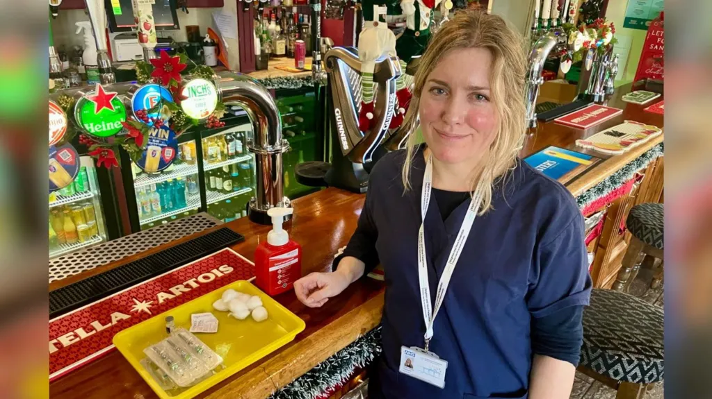 A nurse stands at the bar of a pub with flu vaccination equipment. 