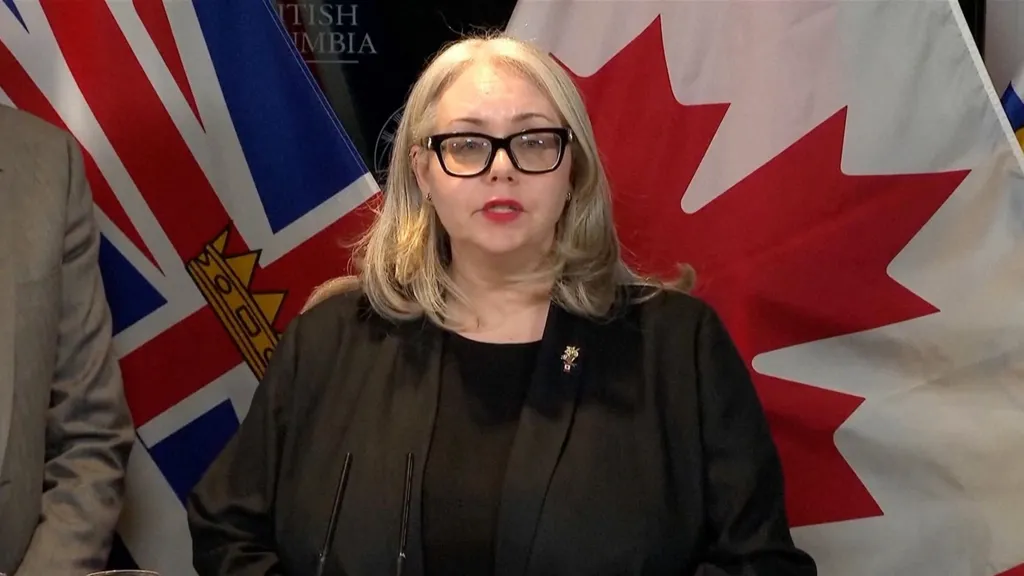 Minister of Public Safety and Solicitor General of British Columbia Nina Krieger. stands wearing a black jacket and top in front of a backdrop of Canadian flags at a press conference