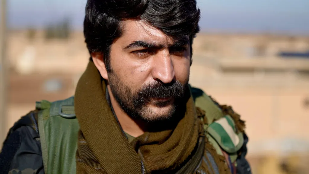 Commander Azad of the Kurdish Forces stares past the camera. He wears a thick black moustache, heavy stubble, and his uniform. 