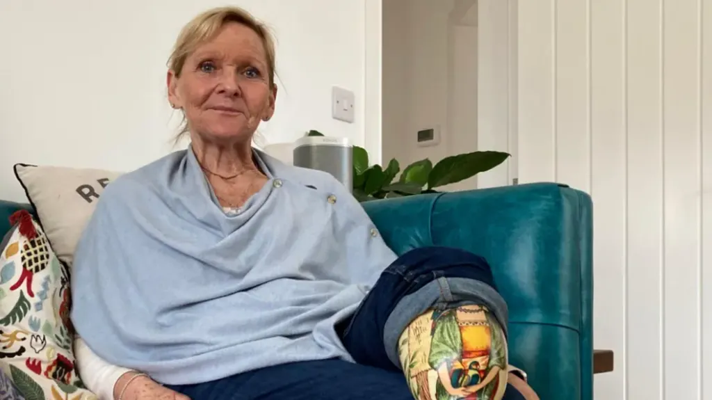 A woman sits on a sofa. She has blonde hair and is wearing a blue top and dark jeans with her prosthetic leg on display. The sofa is covered in floral cushions and colourful blankets. The room has cream walls and a green plant is visible in the background.