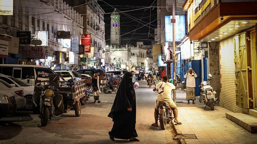 A woman clad in black walks in a street with buildings on both sides , with a man on a motorbike behind her and a mosque minaret in the background
