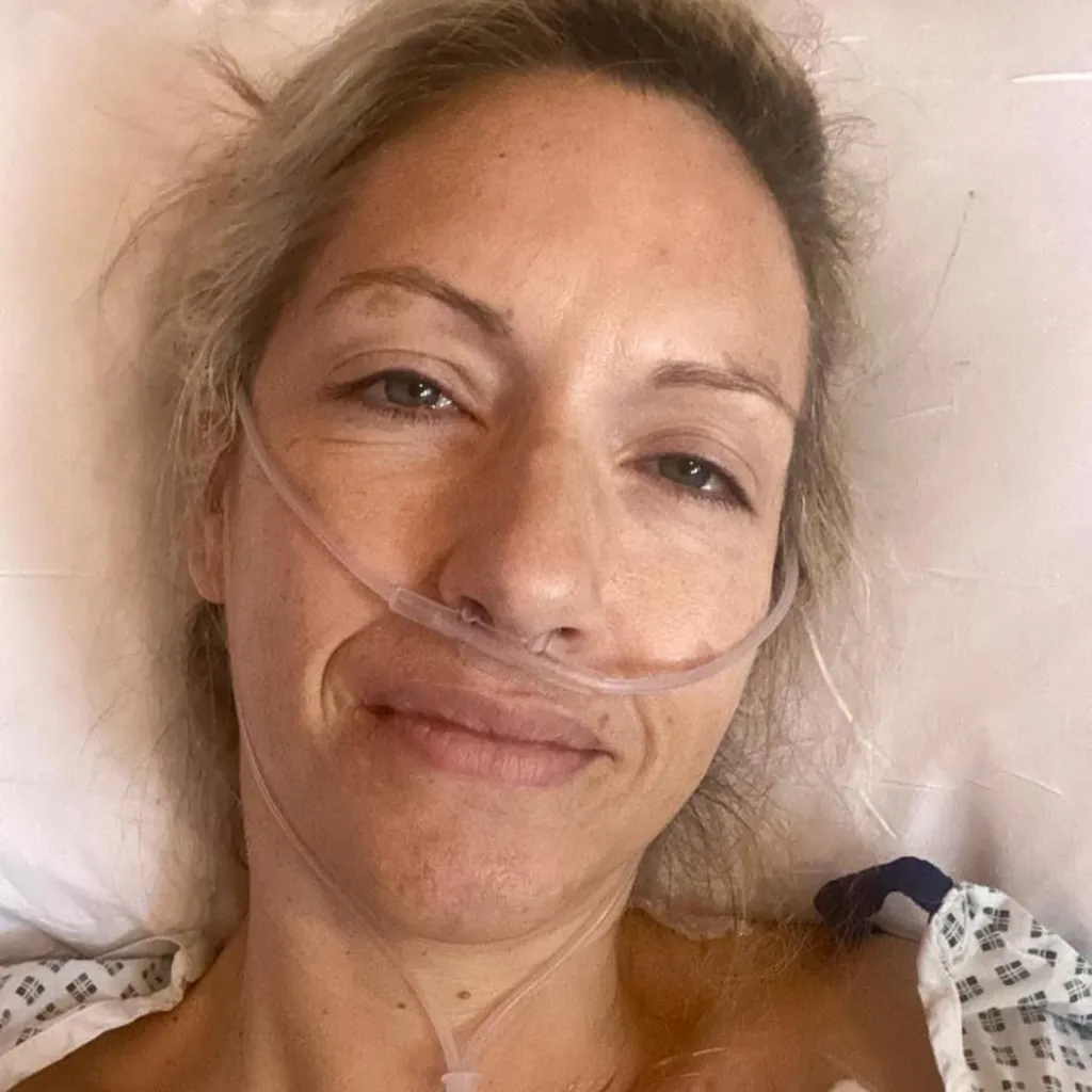 Jessica wearing a hospital gown, lies in a hospital bed with nasal tubes attached. Her blonde hair is tied back. 