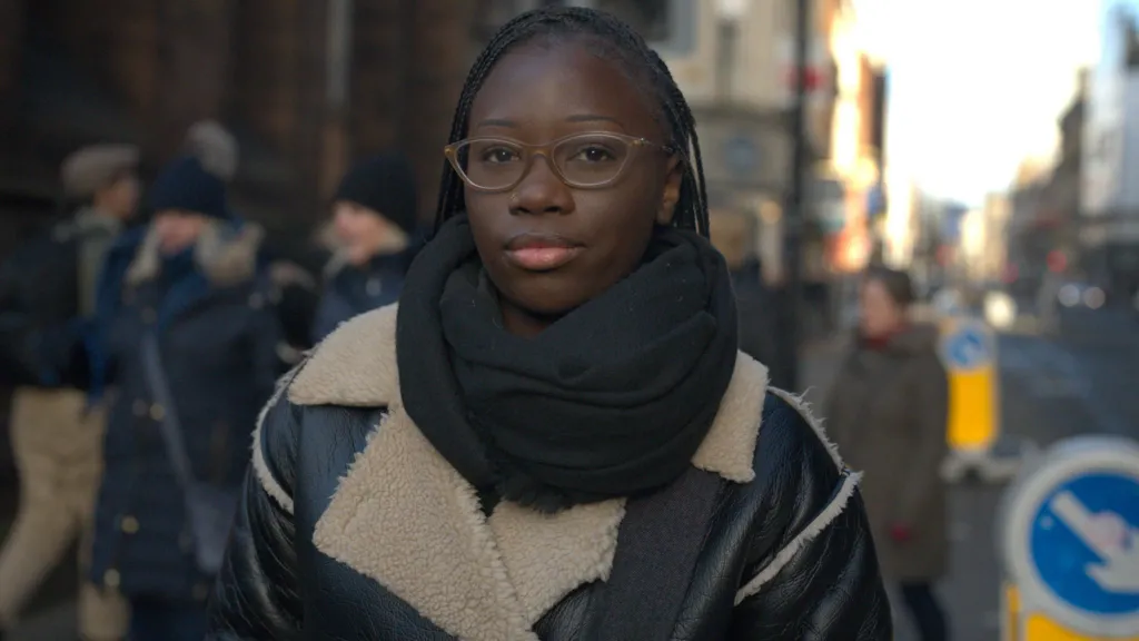 A young black woman wears a black shearling aviator jacket and black scarf. She has braids and looks into the camera wearing glasses. She is standing on a busy road