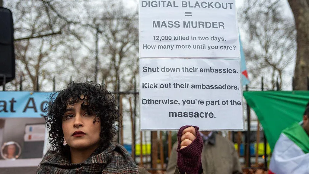 a woman with curly hair holds up a sign that reads "digital blackout = mass murder"