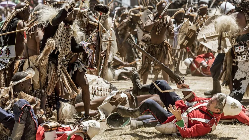 A enactment in 2023 of the Battle of Isandlwana with Zulu warriors with shields and spears bearing down on British soldiers with guns in red jackets and white helmets, several of whom have fallen to the ground. 