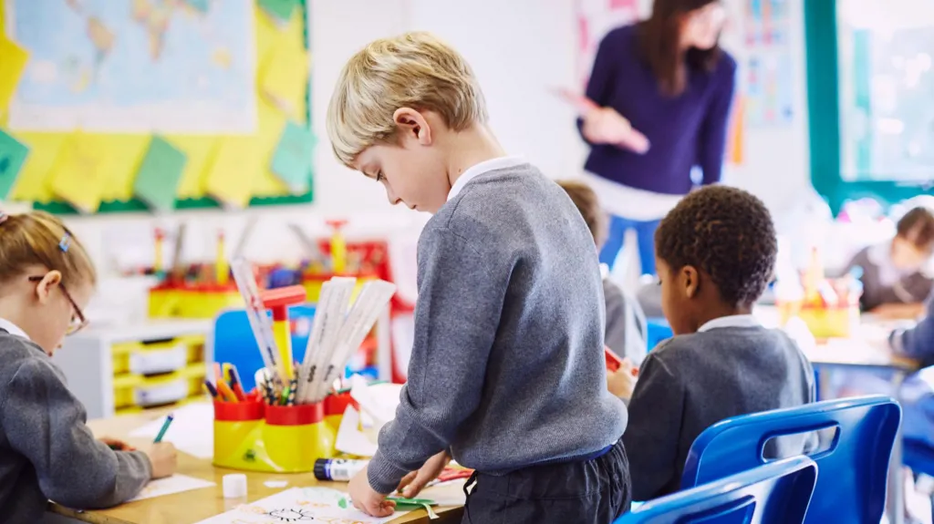 A blonde haired boy in a grey uniform stands at a desk doing a craft. Some other students are sitting at a table. A teacher at the top of the class wearing a purple top looks at some of their work. 