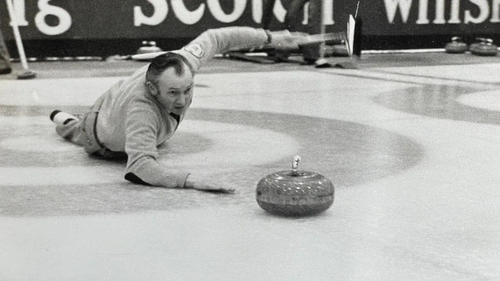 Hammy McMillan lying on his stomach on the ice with his right arm resting on the ice, having just thrown the curling stone- his left arm is held in the air