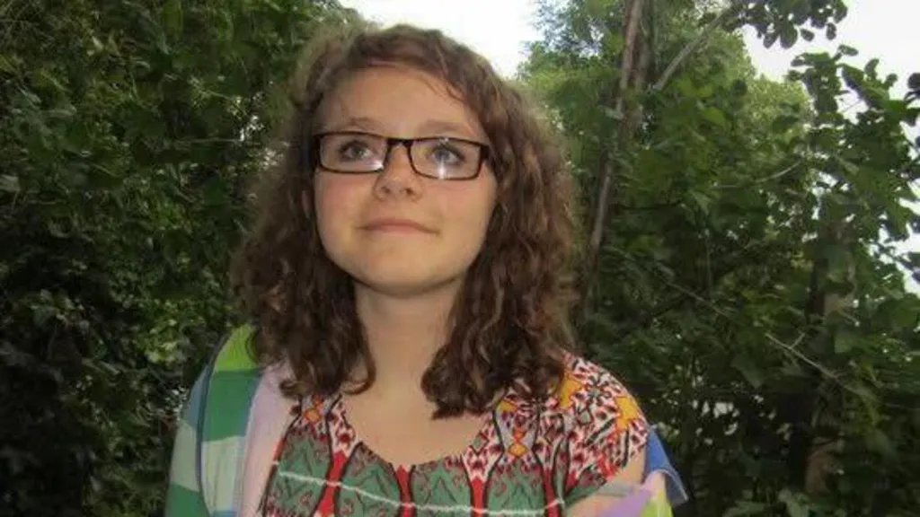 Tessa, who has rectangle framed glasses and dark curly hair. She is wearing a colourful top with green, red, and yellow patterns on it, and a green and blue striped cardigan. Trees can be seen behind her. 
