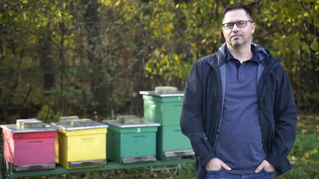 Wearing a blue jacket and blue shirt, Dr Juraj Majtán stands in front of four beehives.
