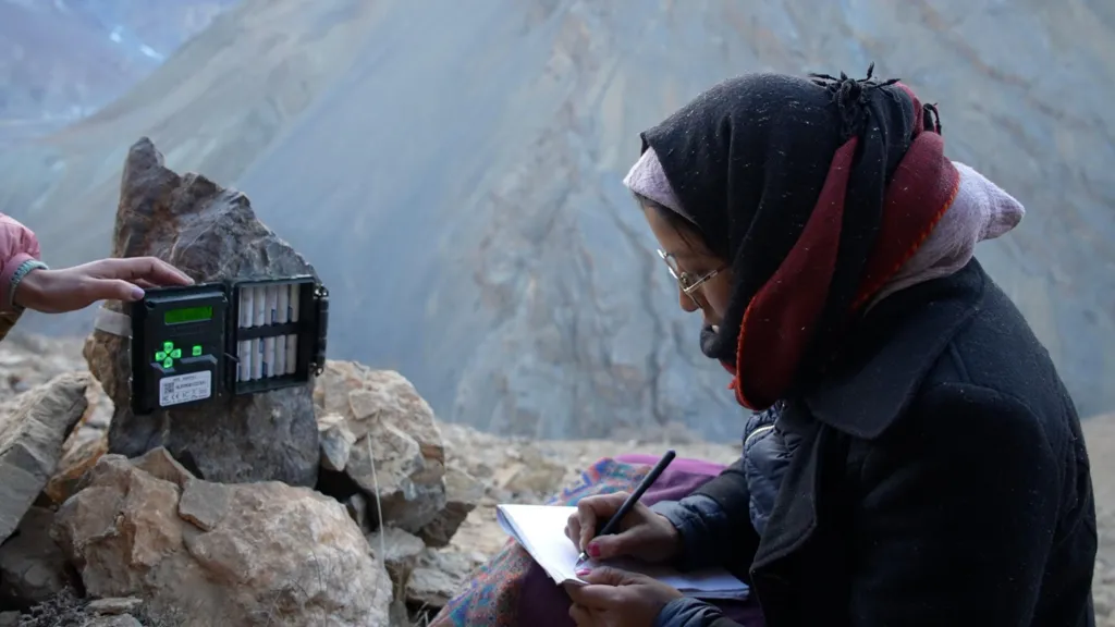 A woman wearing a black and red scarf writes something in her notebook and a camera trap is placed in front of her.