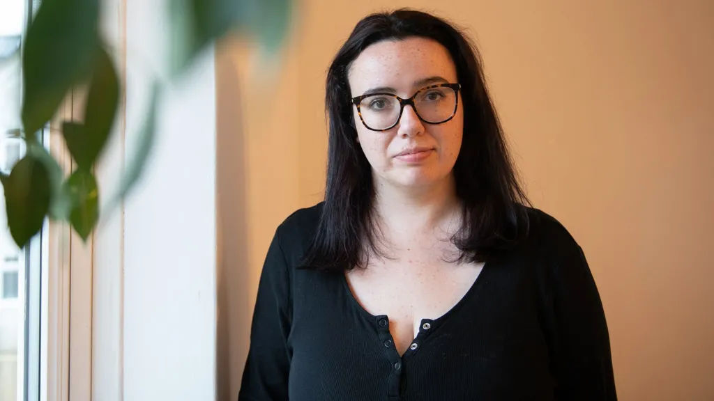 Beth Cox, with long dark hair and glasses stands indoors against a beige wall near a window. Natural light illuminates her face, and large green plant leaves partially obscure the left side of the frame. Buildings are visible through the window behind her.