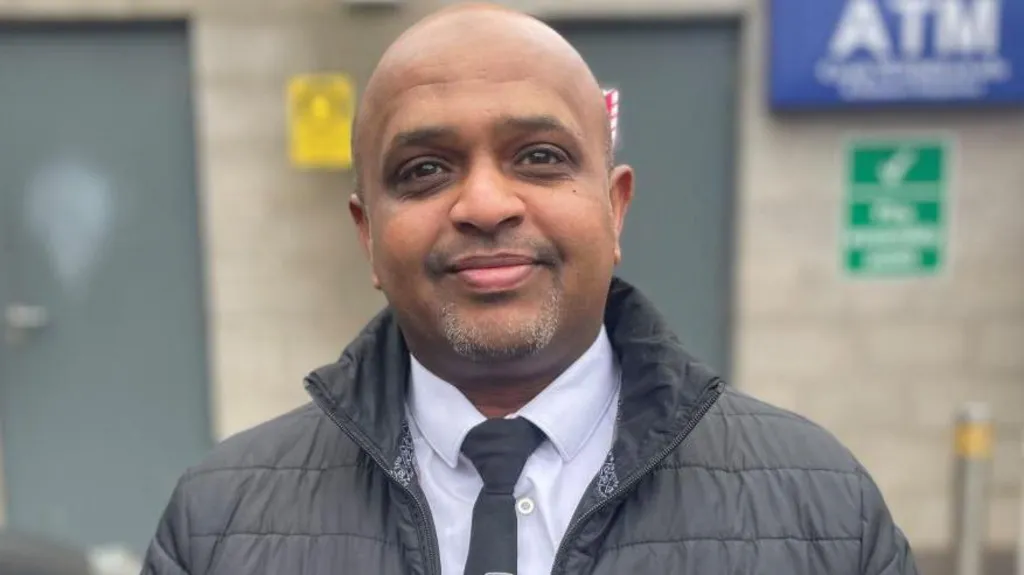 A bald man with grey goatee smiles and looks into the camera. He is wearing a black puffer style coat, white shirt and black tie with the white and yellow 'fonacab' logo.