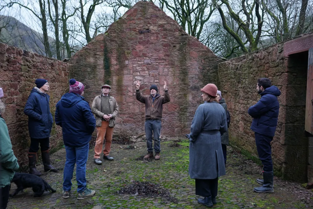 Andrew Goldsworthy stands in derelict farm building that is missing its roof; he is speaking to several people surrounding him 