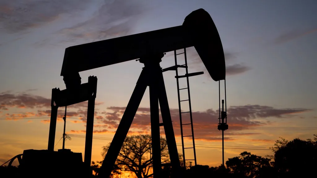 The silhouette of a nodding donkey oil pump against a twilight sky. The outline of a few trees can also be seen. 