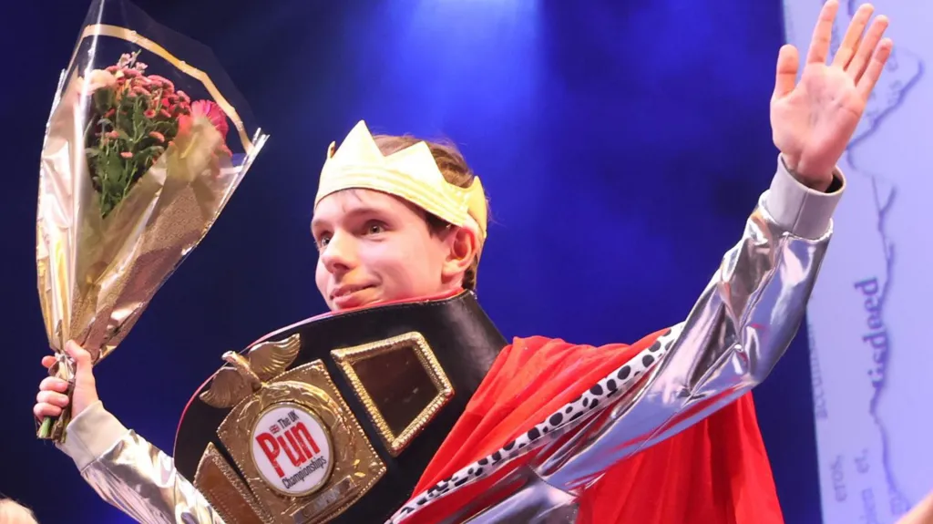 Adam Ernest Pickard with his hands in the air, celebrating winning the UK pun championship with a boxing-style belt around his neck. He's wearing a crown and holding a bunch of flowers.