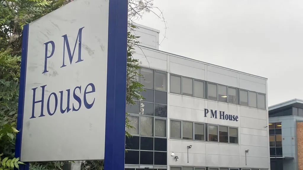 A large office building with the name “PM House” displayed prominently on its exterior. The building has a modern, rectangular design with multiple rows of windows and light grey cladding. In the foreground, a tall sign also reads “PM House” in blue lettering on a white background, framed by a dark blue border. Several cars are parked in front of the building, suggesting a business or office complex. 
