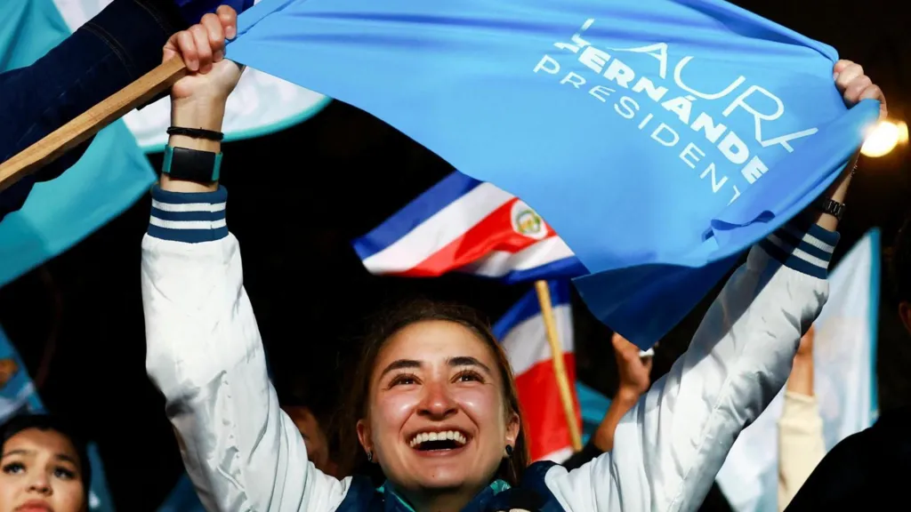 A supporter of Fernández holds up a flag bearing her name and the word "president" in the light blue colour of her party. The young woman is smiling broadly. Behind her, other supporters can be seen also waving flags. 