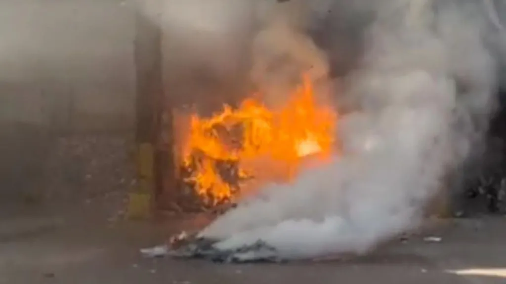 A fire rages in a recycling facility. Plumes of smoke rise in the foreground, in front of a pile of material burning with bright orange flames.