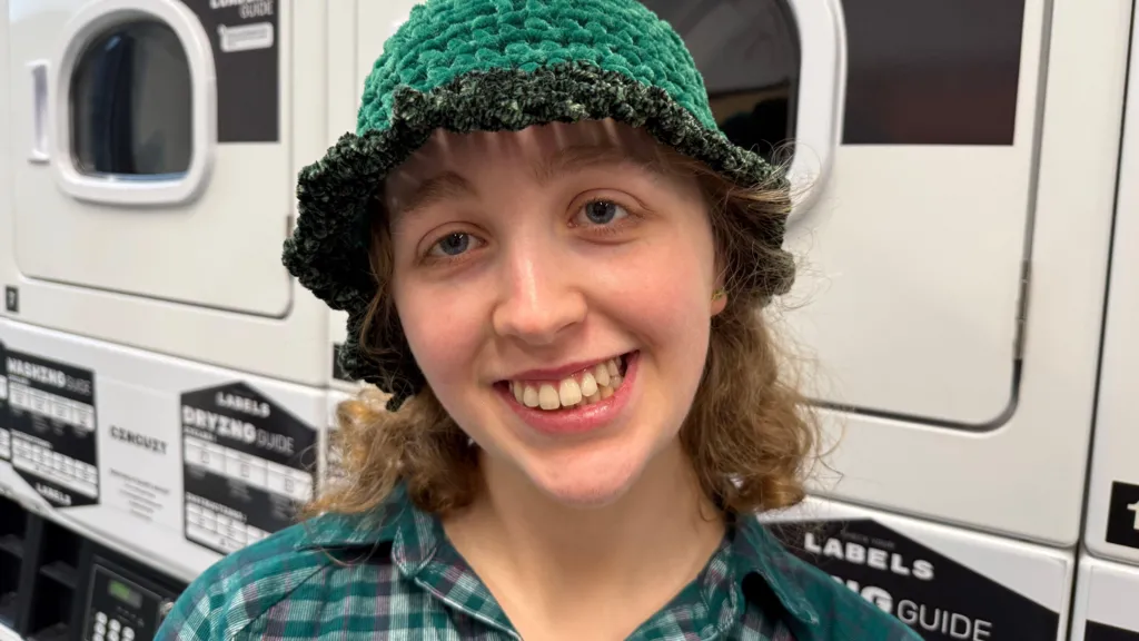 Cathy is a disabled student. She is standing in front of the washing machines in a laundry room on campus at the University of York. Cathy is smiling and wearing a green checkered shirt alongside a green hat covering her curly hair. 