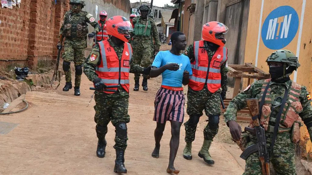 A man in blue jersey and stripped shorts is accosted by security forces in military uniform in Kampala