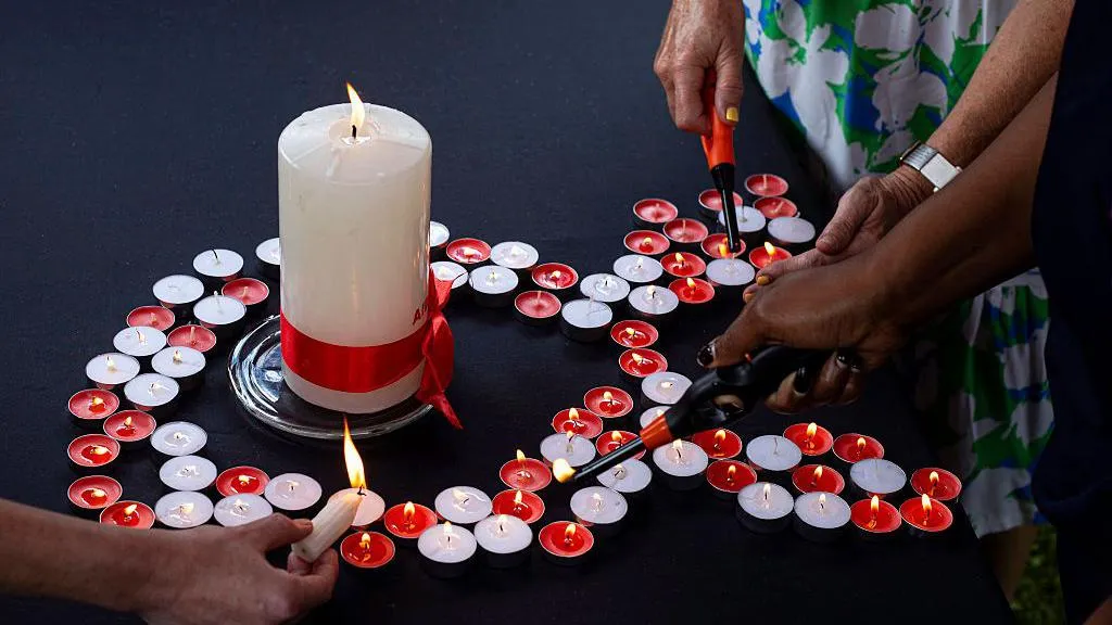 Small red and white candles are arranged in the shape of an Aids ribbon. A large white candle is in the centre. The candles are being lit by some disembodied hands. 