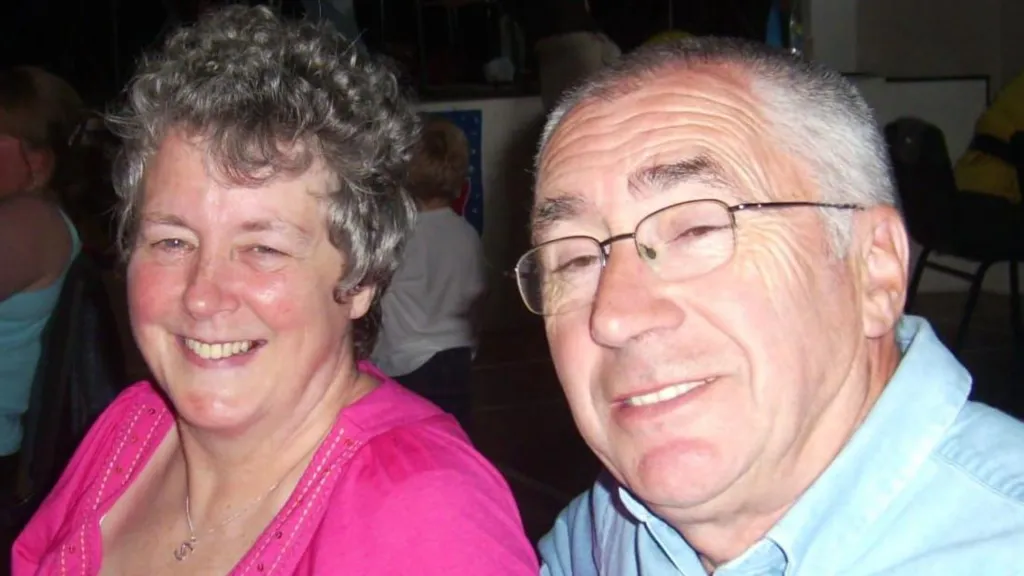 A couple pictured in a room with others in the background. They are both smiling. The woman is wearing a pink scoop-necked t-shirt and has light curly short hair. The man is in a pale blue shirt and wire-framed glasses. He has white hair swept back from his forehead.