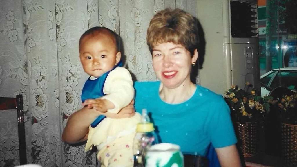 A woman in a blue top smiles at the camera. She has short fair hair. A baby wearing a yellow top and a blue bib, being held by the woman, looks at the camera too.