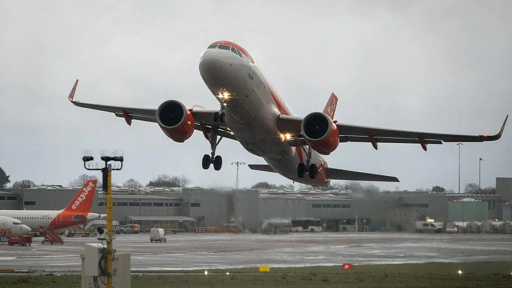 An EasyJet aircraft in the air as it leaves the runway at Bristol Airport on an overcast day. Gray hangers can be seen in the background, along with maintenance vehicles and buses. The rear orange tail of an EasyJet plane can be seen to the left of the frame.