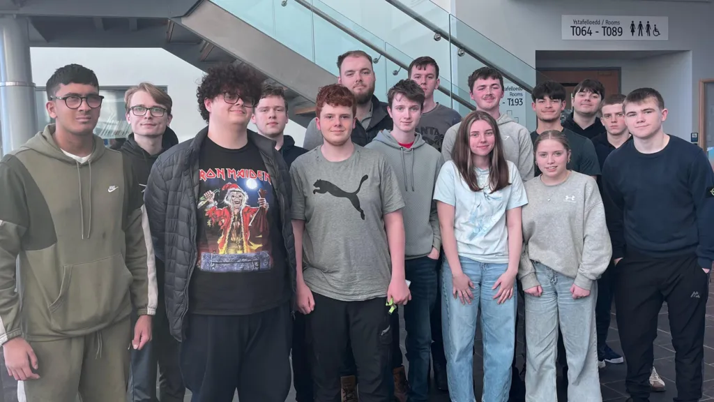 A group of 15 students, mostly wearing hoodies, jeans, t-shirts and jogging bottoms are lined up and looking directly at the camera in front of a flight of glass and metal stairs in a foyer. 