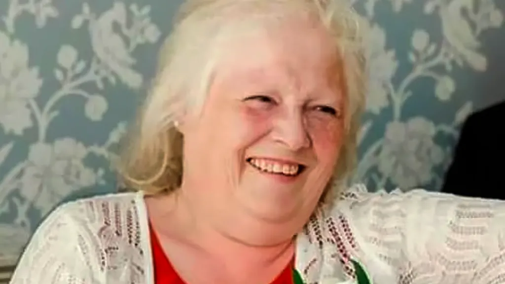 Esther Martin is wearing a red top and a white cardigan with a flower pinned to it. She is smiling.