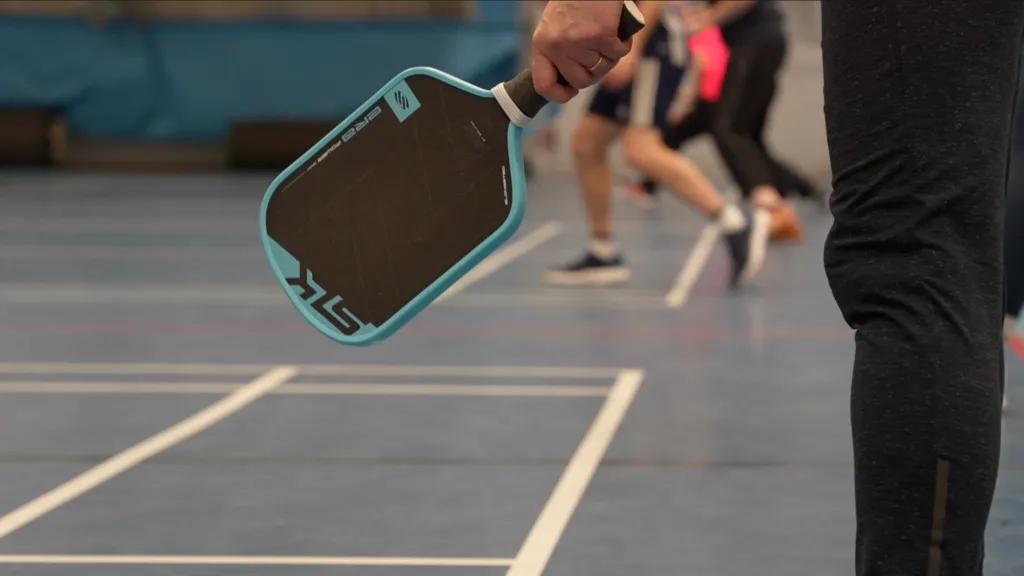 A hand holding a pickleball paddle, face open. The paddle has a light blue rim, and a black surface where the polymer ball strikes. There are players, mid-match, in the background. 
