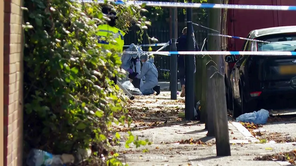 Forensic investigators in white protective suits examine a cordoned‑off residential street while a police officer in a high‑visibility jacket stands nearby. Blue-and-white “Police Do Not Cross” tape blocks the area, with a parked car and fallen leaves visible along the pavement.