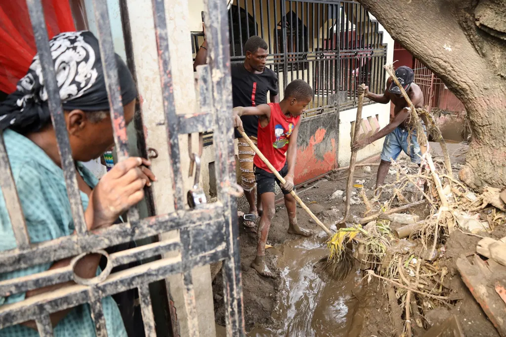 People clean up debris in front of a flooded house as a family member looks on, in Petit Goave, Haiti on 31 October 2025.