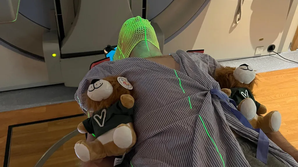 Anthony Perriam is lying in a hospital bad with two teddy bears by his side. He is wearing a mask as he is about to be scanned for cancer.