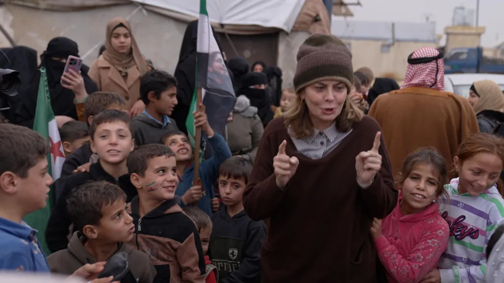 Kabawat in a brown jumper and woollen hat, surrounded by about 10 children, several of whom are looking up at her. Some are smiling. She is speaking and gesturing with her two forefingers outstretched. Tents in the camp for displaced people can be seen behind her. 