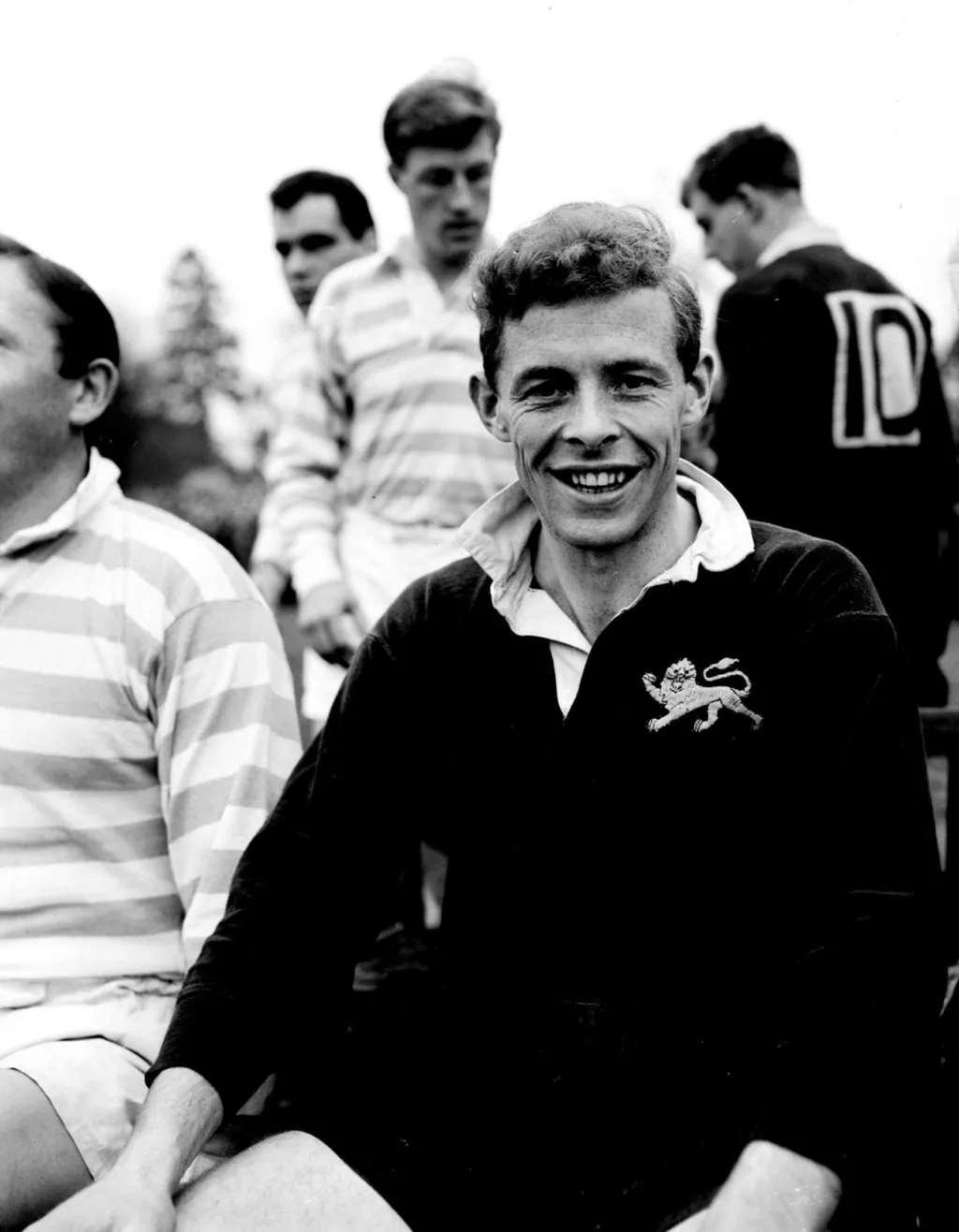 A black and white photograph of Arthur Smith as a young man. He is smiling broadly and wearing a dark-coloured rugby top. he is surrounded by others in similar clothes. 