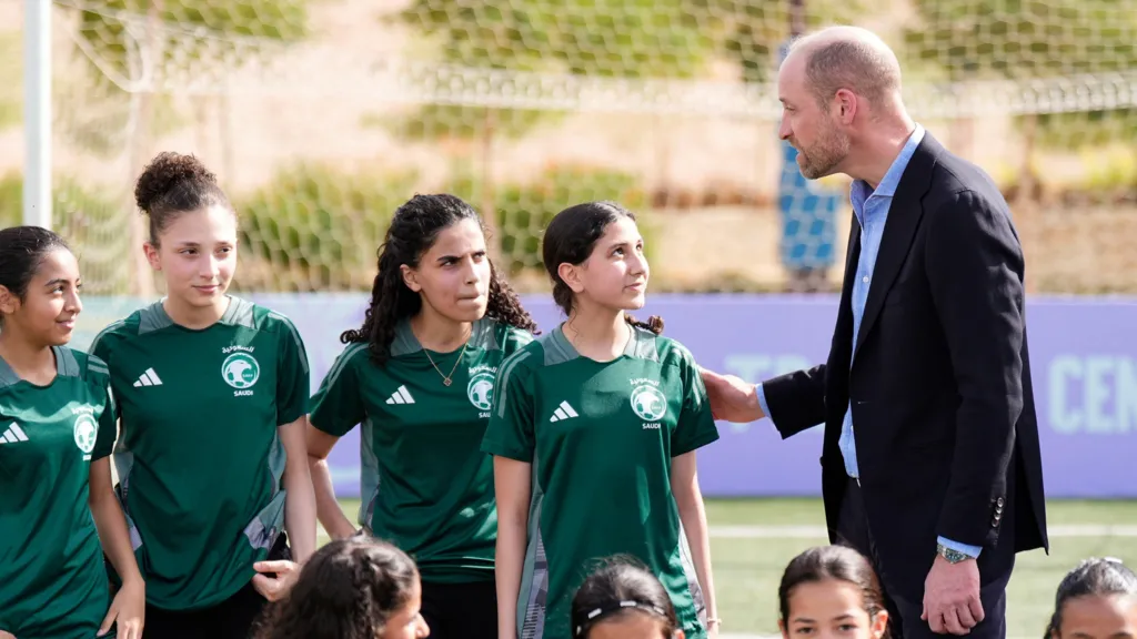 Prince William talks to a group of young girls in green football kits.