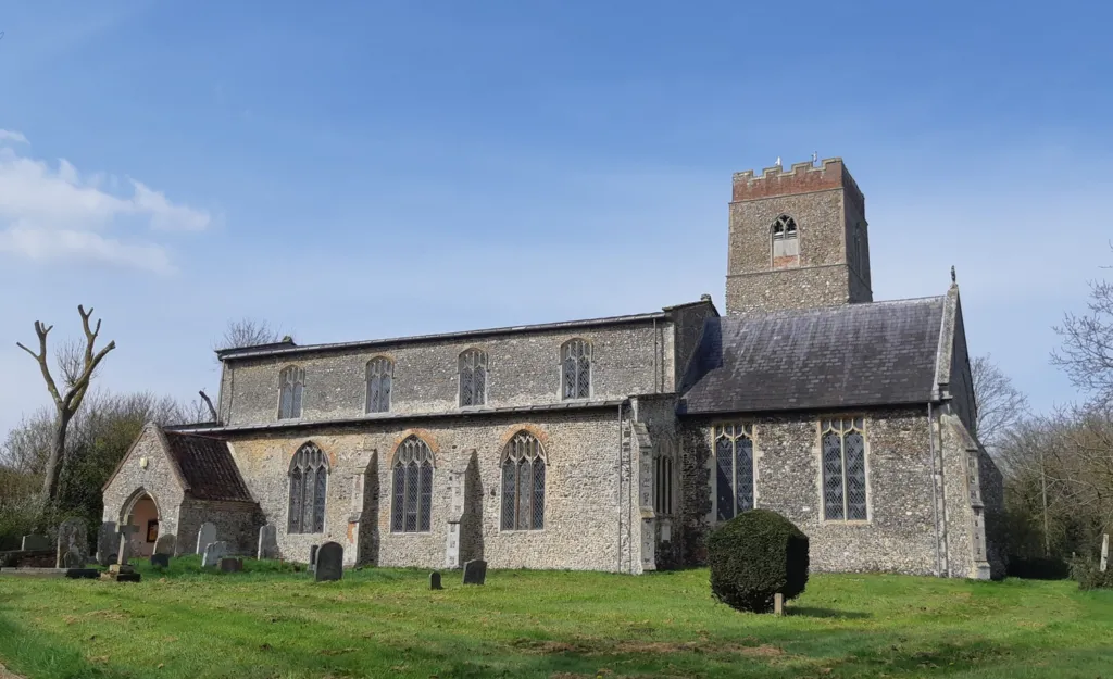 Exterior of Guestwick Church in Norfolk taken on a clear winter's day. The view is of the side of the church - it's walls are made from grey flint stone, and it has arched stained glass windows and a tower. In the foreground there are some gravestones on a green lawn. 