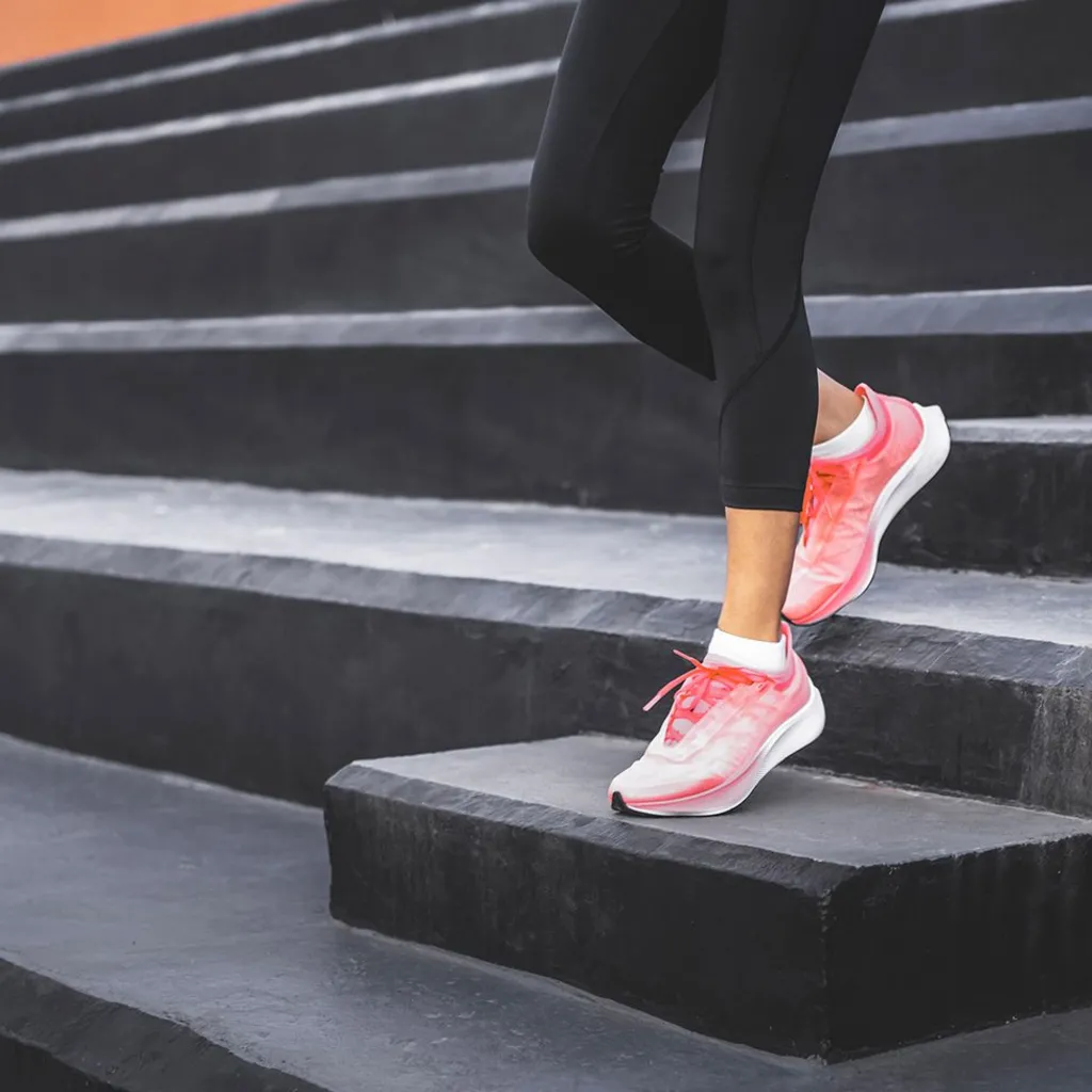 A woman wearing black leggings and pink athletic shoes, standing on a set of black steps. One foot is placed on a higher step while the other remains on a lower one, suggesting an exercise or stretching.